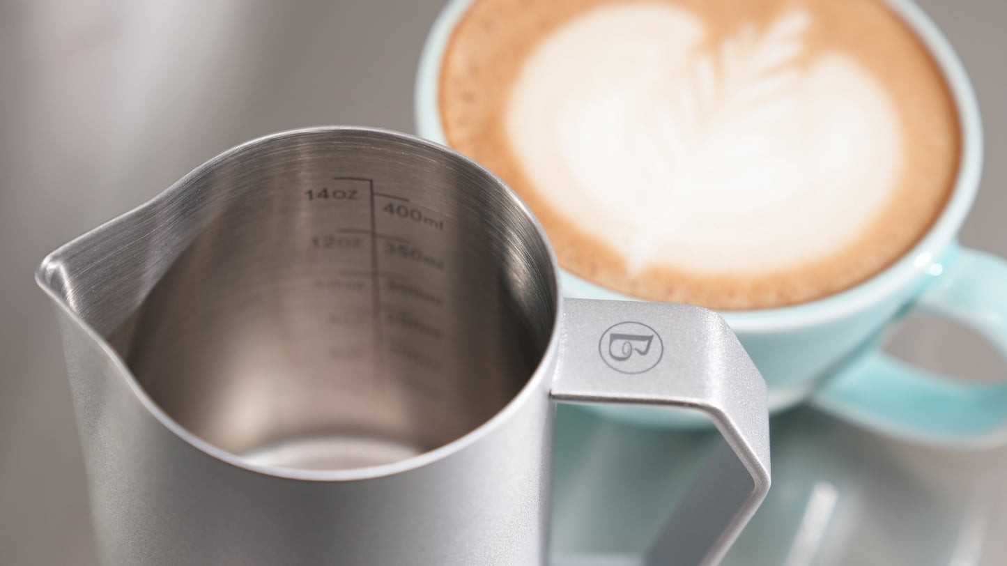 Metal pitcher with measurement markings next to a cup of coffee